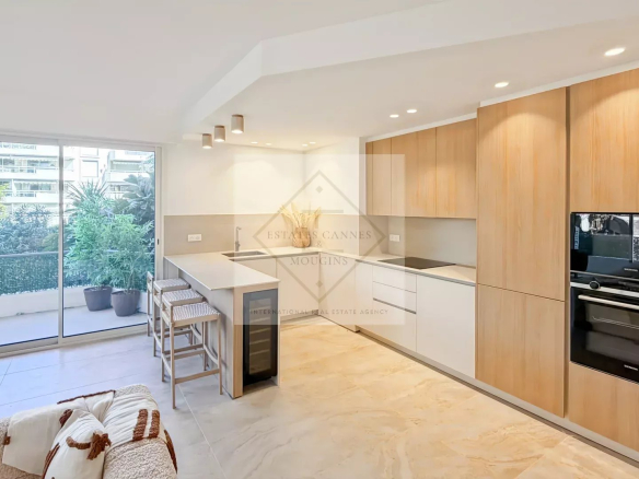 Modern kitchen with white countertops and light wood cabinets, island seating, and a sliding glass door to a plant-filled balcony outside.