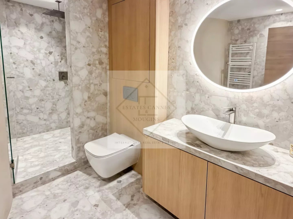 Modern marble bathroom with glass shower, wall-hung toilet, wood vanity, oval vessel sink, and a round backlit mirror.