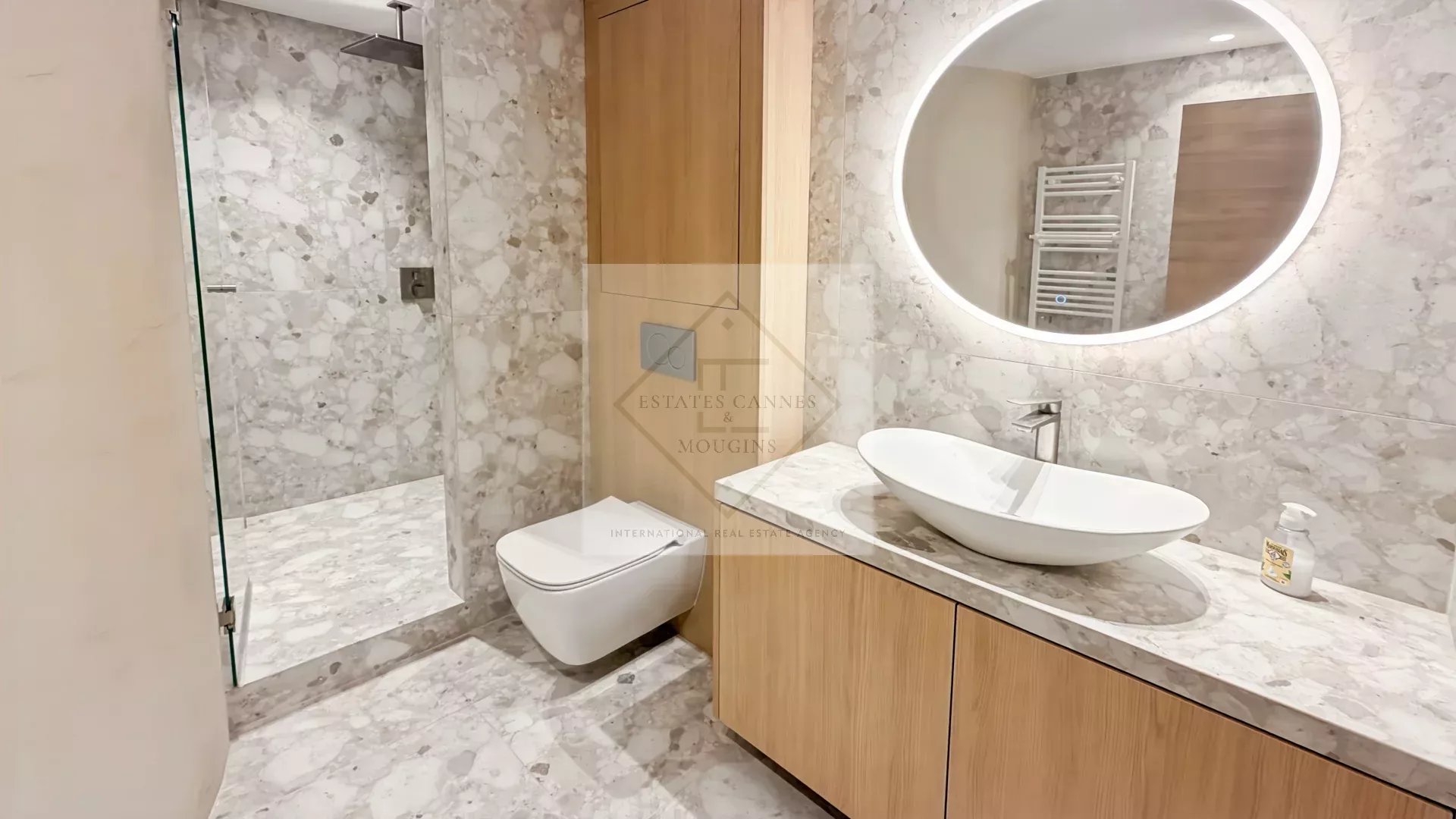 Modern marble bathroom with glass shower, wall-hung toilet, wood vanity, oval vessel sink, and a round backlit mirror.