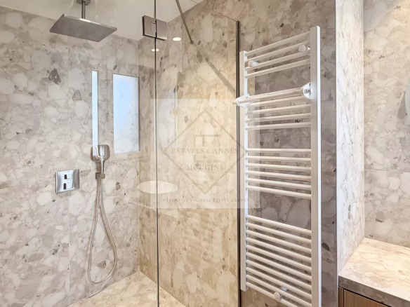 Modern marble bathroom with a glass-walled shower, rainfall and handheld shower fixtures, and a white wall-mounted towel warmer next to a round mirror.