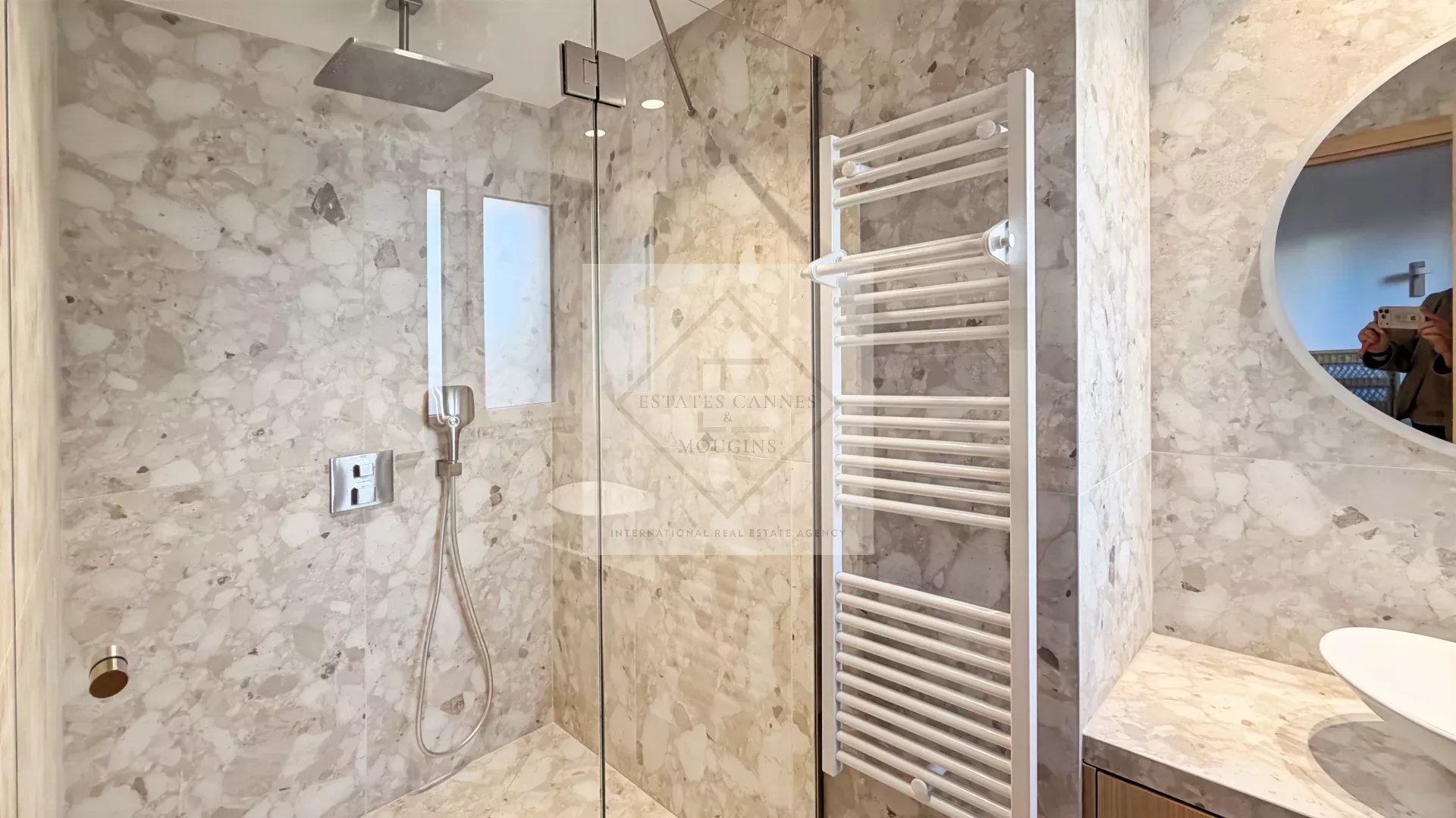Modern marble bathroom with a glass-walled shower, rainfall and handheld shower fixtures, and a white wall-mounted towel warmer next to a round mirror.
