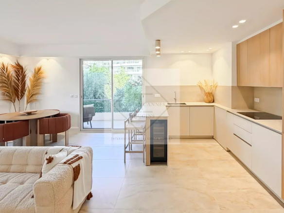 Bright open-plan living area with a beige sofa, round dining table and pink upholstered chairs, and a modern kitchen with wooden cabinets and light flooring.