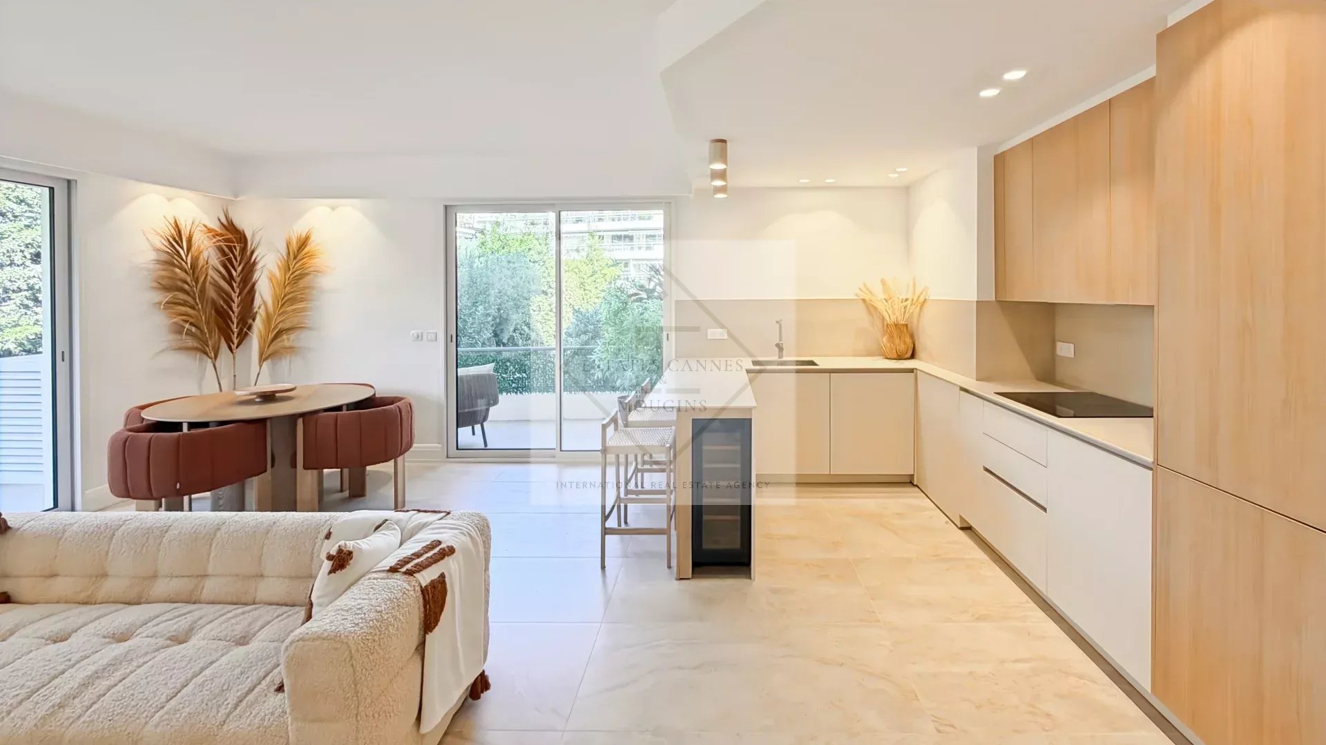 Bright open-plan living area with a beige sofa, round dining table and pink upholstered chairs, and a modern kitchen with wooden cabinets and light flooring.