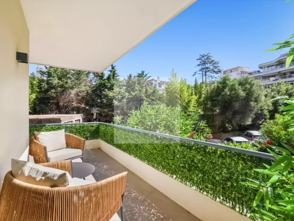Sunny balcony with two wicker chairs and a small table, overlooking a lush hedge and trees.