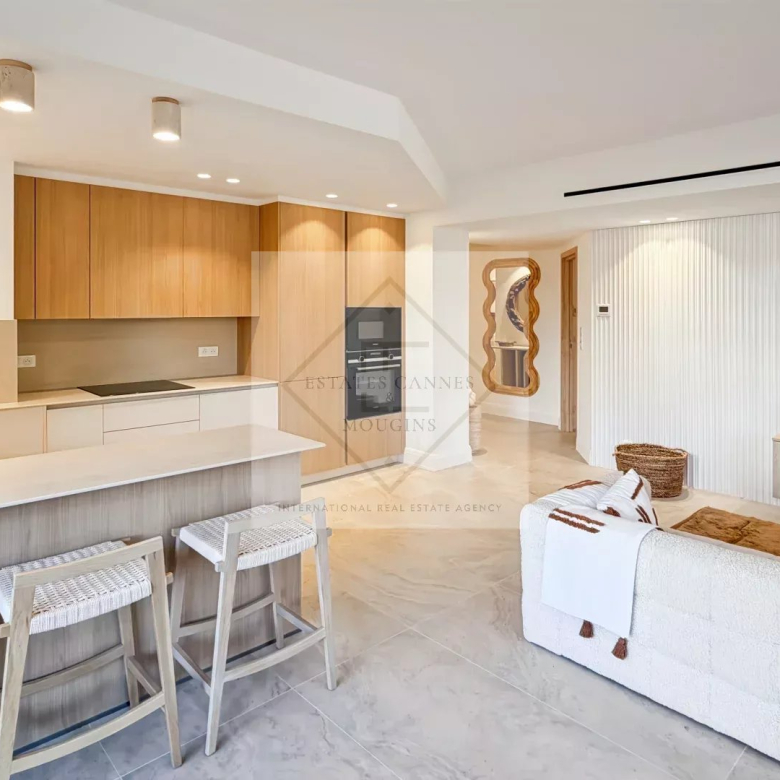 Open-concept living space with a white kitchen island, light wood cabinets, and three woven-bar stools; airy and modern.