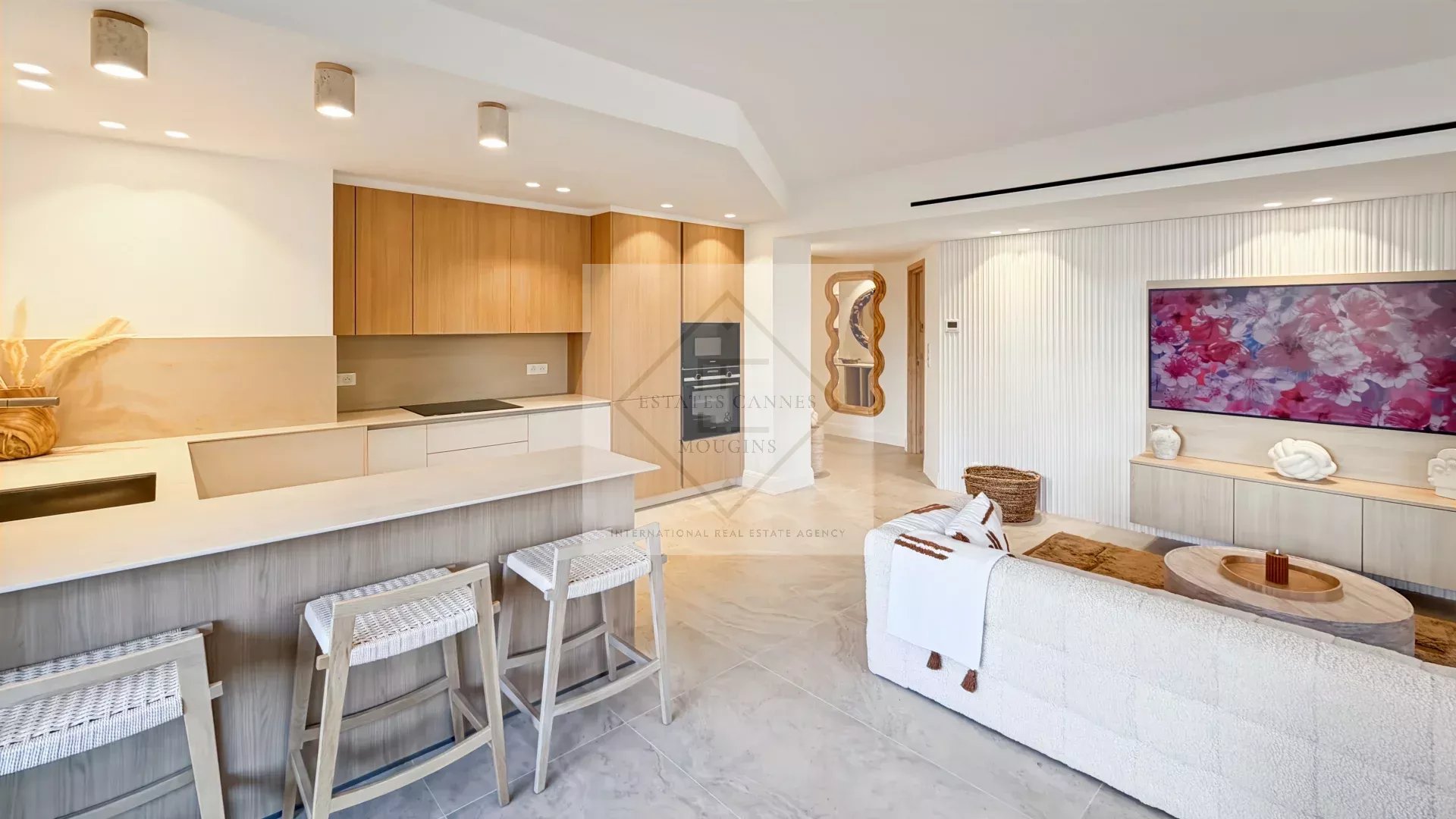 Open-concept living space with a white kitchen island, light wood cabinets, and three woven-bar stools; airy and modern.