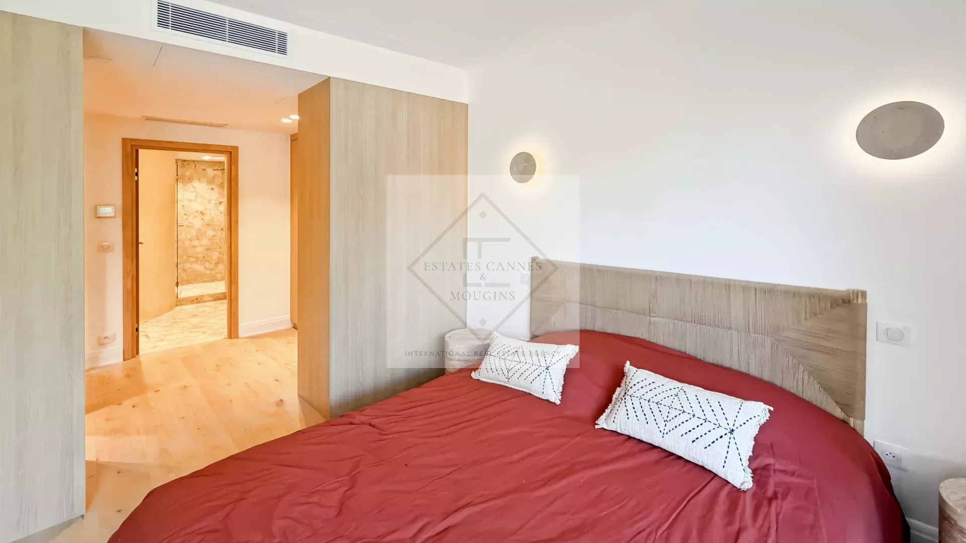 Bright bedroom with a red bedspread, white pillows, and a light wood headboard; doorway to a hallway on the left.