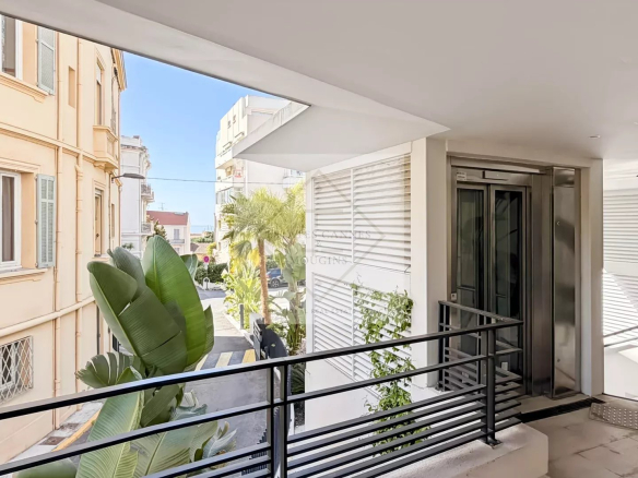 Modern balcony or walkway with black metal railing, next to an elevator door and white louvers, overlooking a sunlit street with palm plants in view.