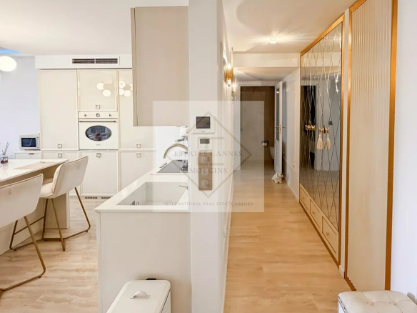 Bright white kitchen with an island, built-in oven, and beige bar stools; leading to a hallway with a large mirrored wardrobe.