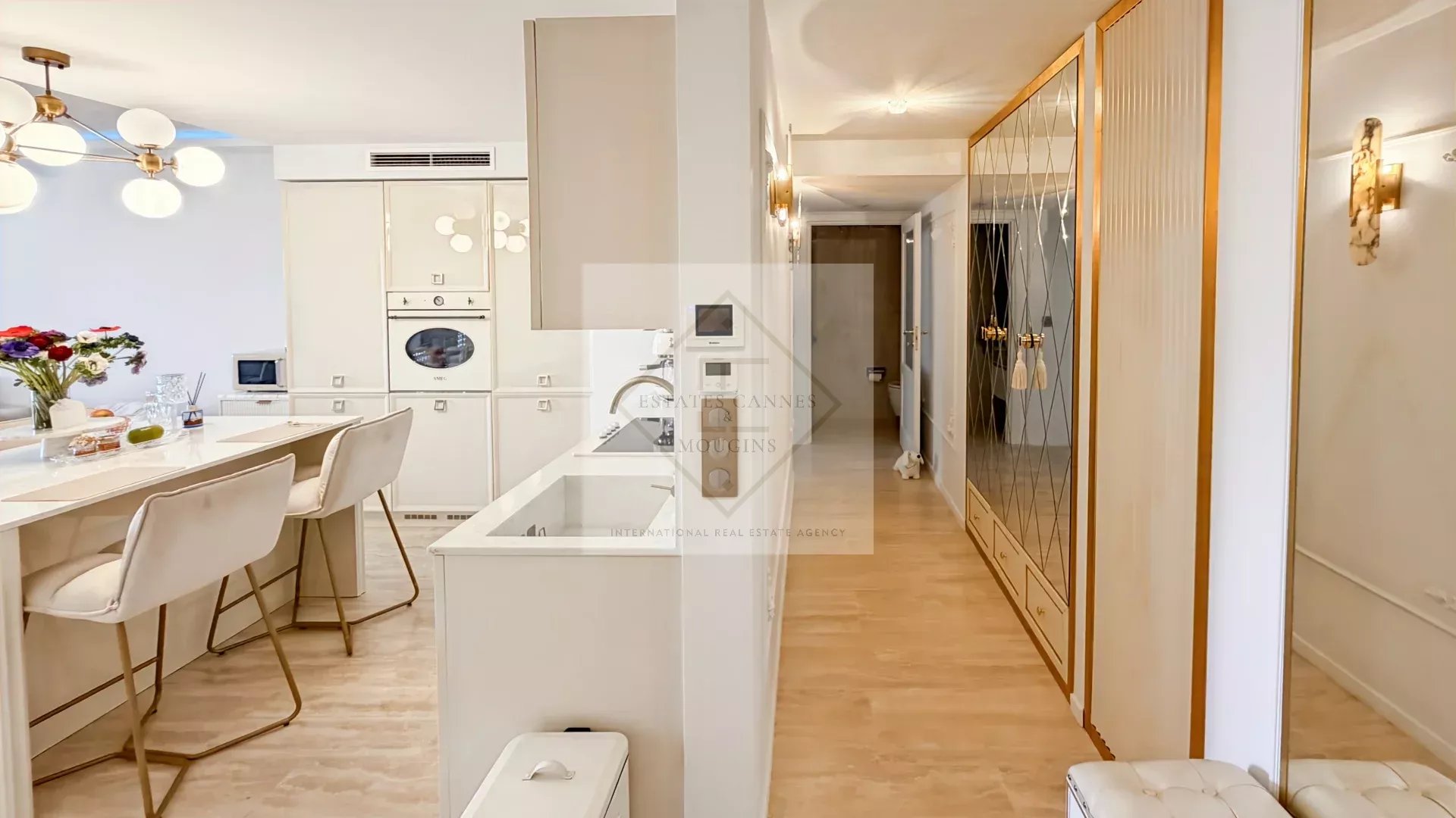 Bright white kitchen with an island, built-in oven, and beige bar stools; leading to a hallway with a large mirrored wardrobe.