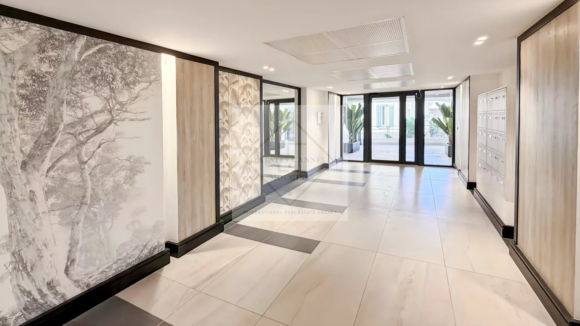 Modern residential lobby with glass entry doors, mailboxes on the right, and decorative wall panels; greenery visible outside.