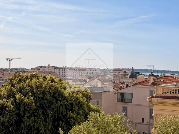 Cityscape of a coastal town with red-tiled rooftops, a construction crane in the distance, and greenery in the foreground under a clear blue sky.