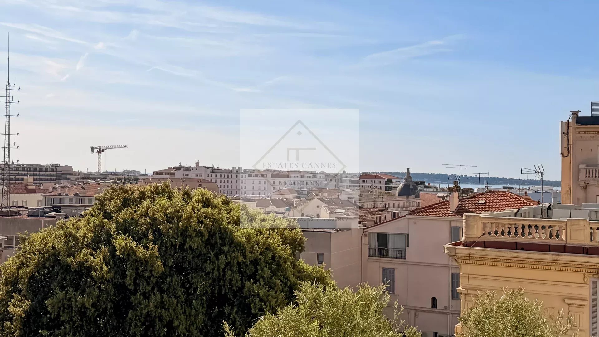 Cityscape of a coastal town with red-tiled rooftops, a construction crane in the distance, and greenery in the foreground under a clear blue sky.