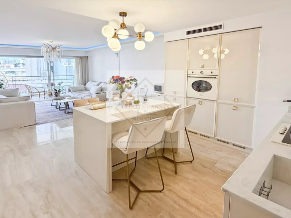 Open-plan kitchen and living room with white cabinetry and marble floors; island seating with two chairs, bouquet on the island, and a balcony view.