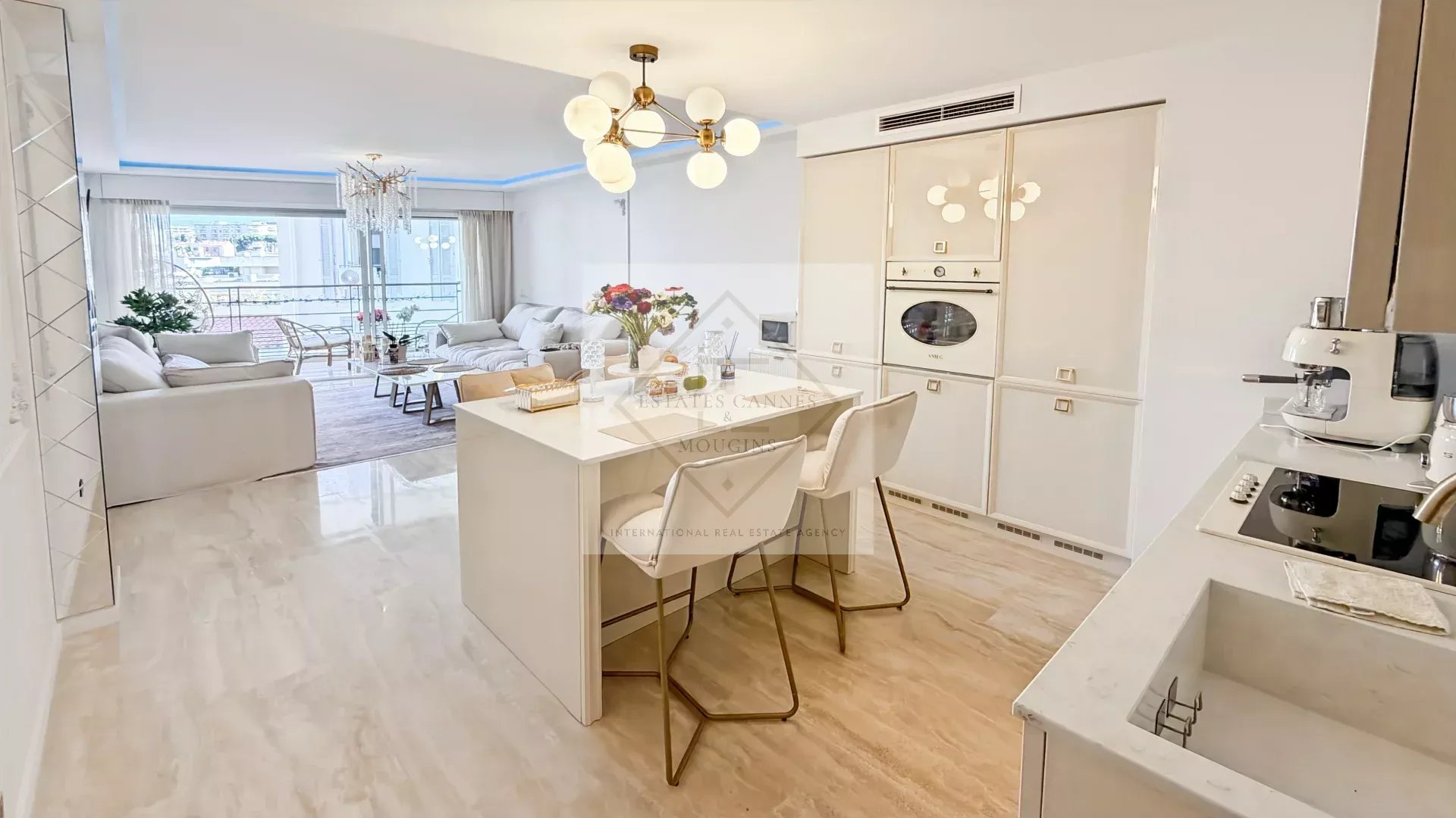 Open-plan kitchen and living room with white cabinetry and marble floors; island seating with two chairs, bouquet on the island, and a balcony view.