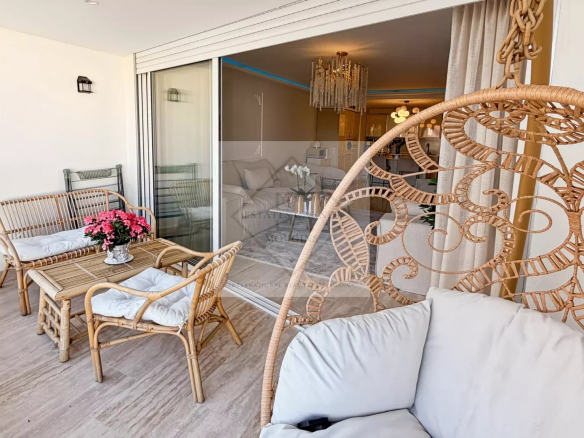 Balcony seating area with rattan chairs and a small table, pink flowers in a pot at the center, sliding glass door to the living room behind.