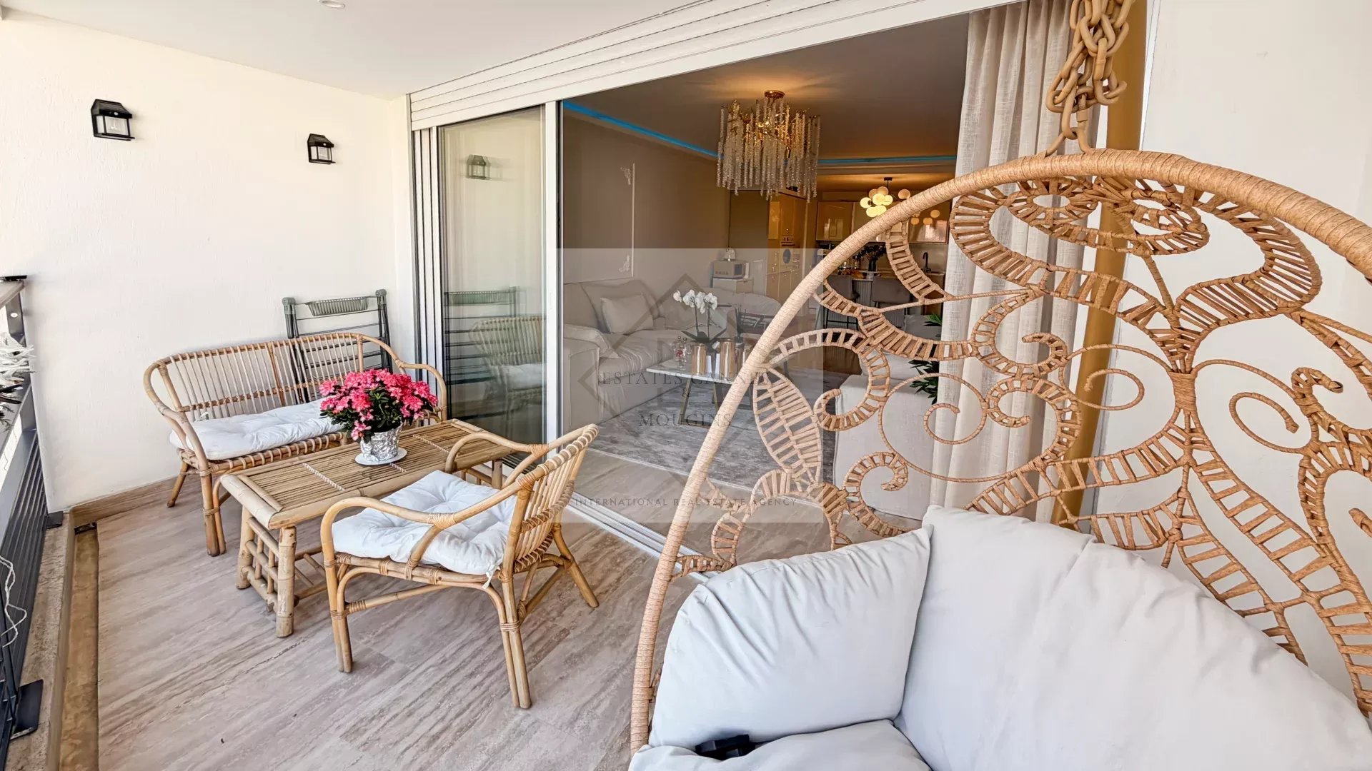 Balcony seating area with rattan chairs and a small table, pink flowers in a pot at the center, sliding glass door to the living room behind.