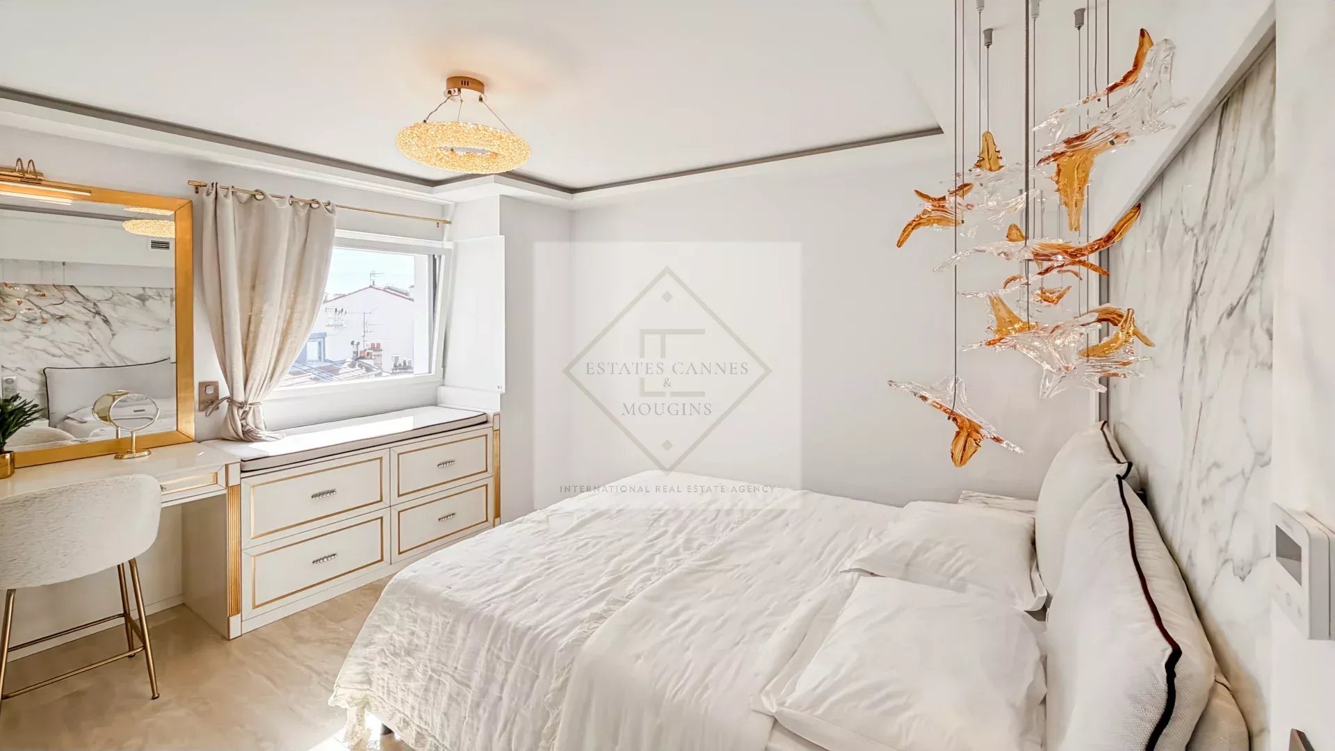 Luxurious white bedroom with gold-accent furniture, marble wall, and a white bed with plush pillows under an orange glass chandelier.