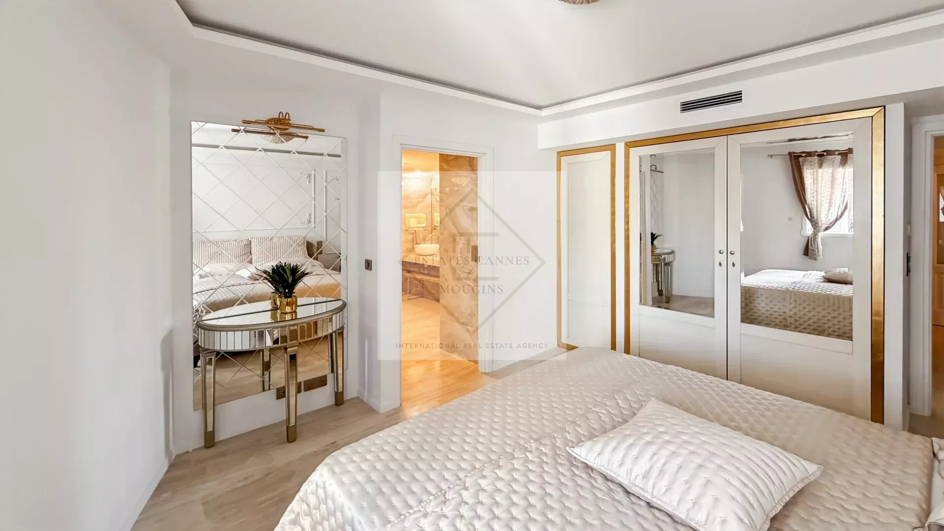 Bright master bedroom with white decor, gold-framed mirrors, and a round glass console table with a plant beside tufted bedspreads.