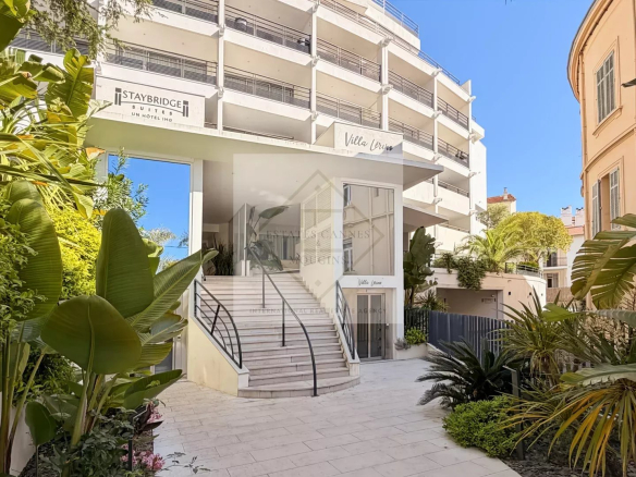 Hotel entrance with white modern facade, curved stairs, and lush tropical plants on a sunny day; Staybridge Suites sign visible.