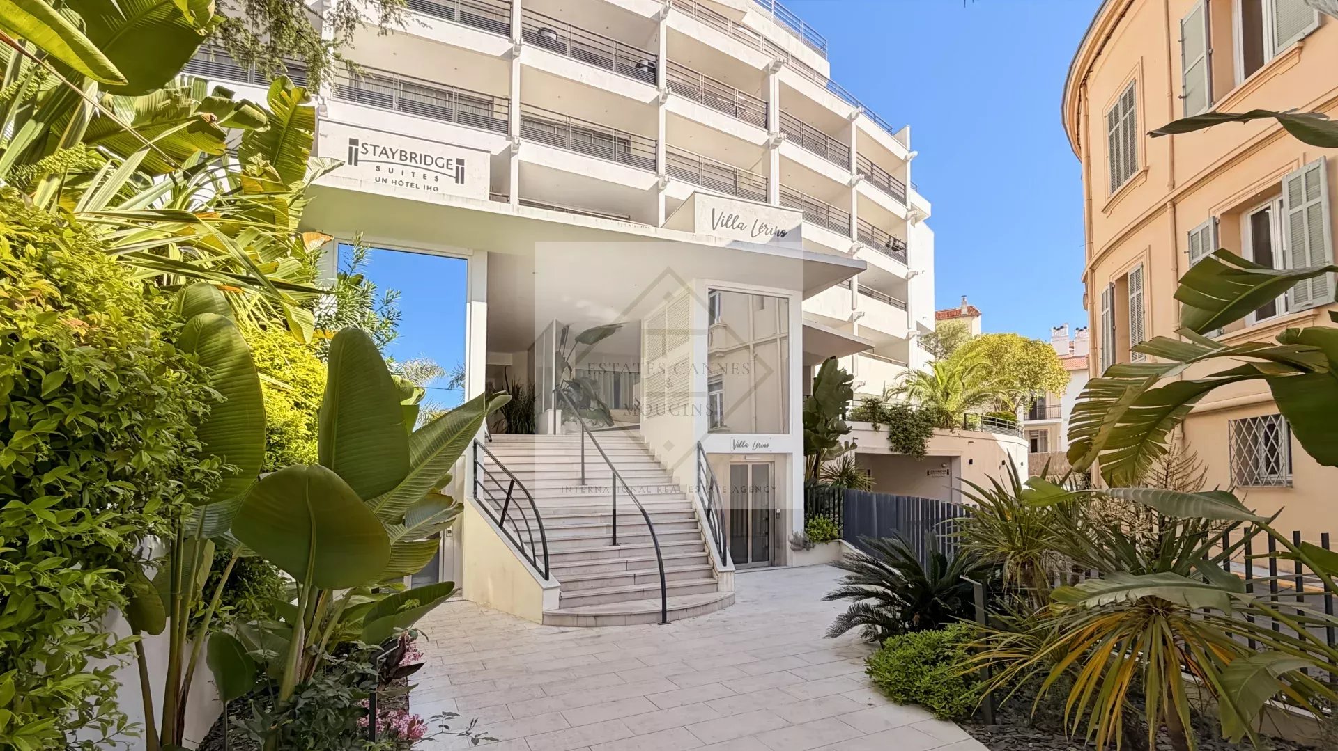 Hotel entrance with white modern facade, curved stairs, and lush tropical plants on a sunny day; Staybridge Suites sign visible.