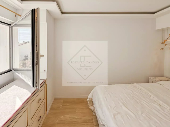 Bright bedroom with a large open window, white walls, and a white bed with a marble headboard; built-in drawers run along the window wall. Outside shows trees and rooftops.] ,