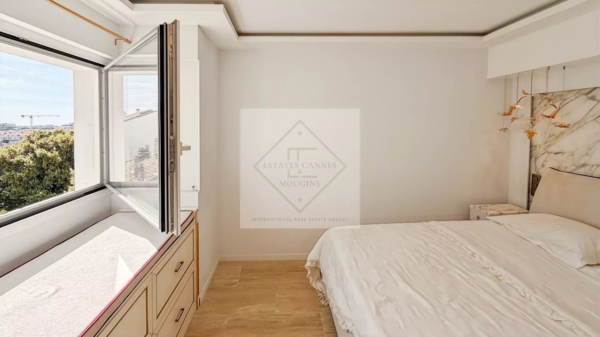 Bright bedroom with a large open window, white walls, and a white bed with a marble headboard; built-in drawers run along the window wall. Outside shows trees and rooftops.] ,