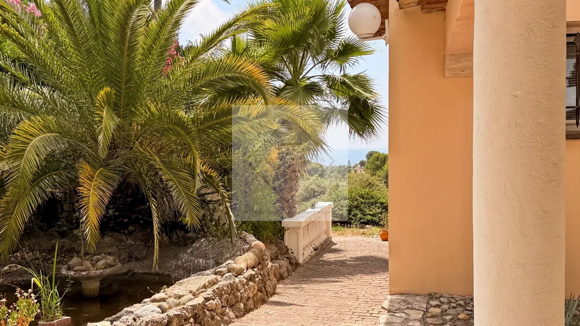 Driveway leading to a villa with sea views, surrounded by palm trees and greenery in Antibes.
