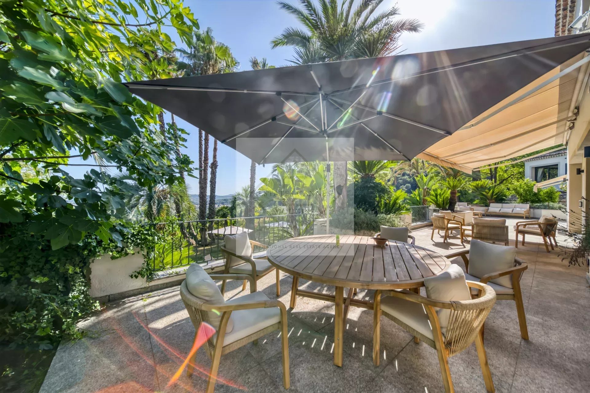 Terrasse ensoleillée avec table en bois et chaises, entourée de verdure et de palmiers.