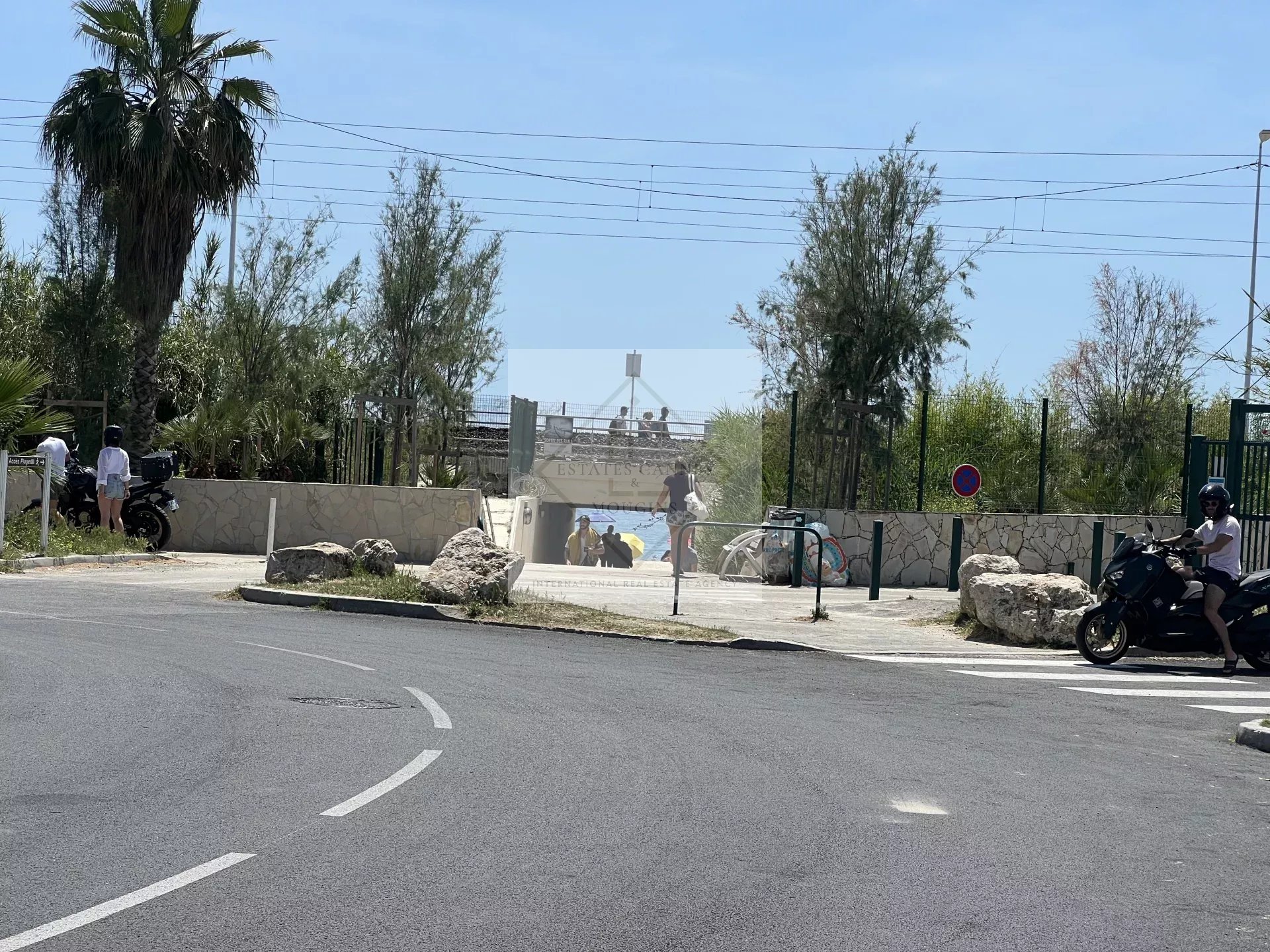 Accès à la plage depuis une rue à Juan-les-Pins, avec des palmiers et des personnes se dirigeant vers la mer.