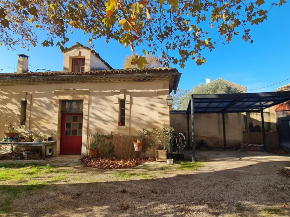 Sunny courtyard scene: beige house with a red front door, potted plants on a table, and a metal-framed covered area to the right under a bright blue sky.
