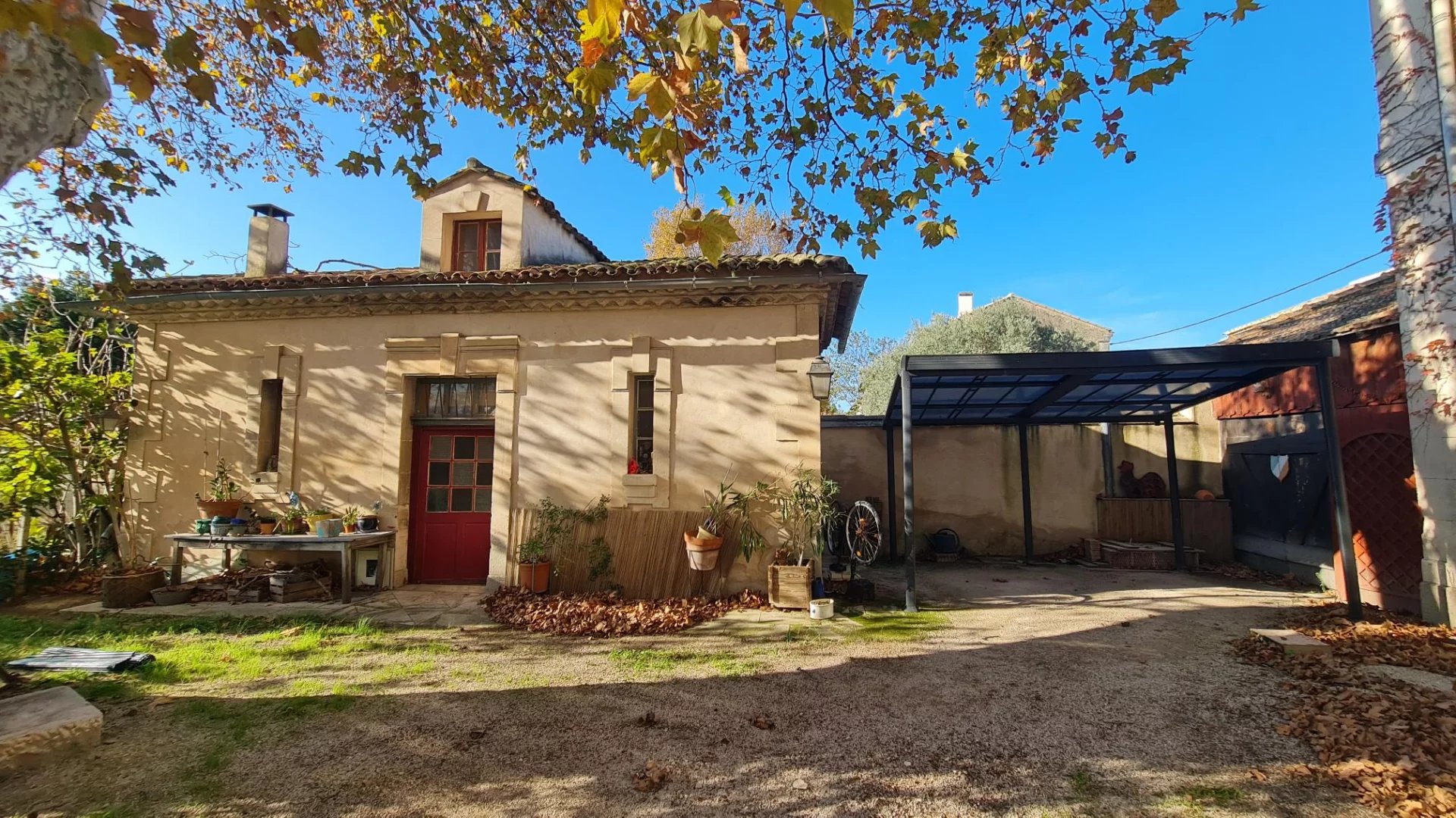 Sunny courtyard scene: beige house with a red front door, potted plants on a table, and a metal-framed covered area to the right under a bright blue sky.