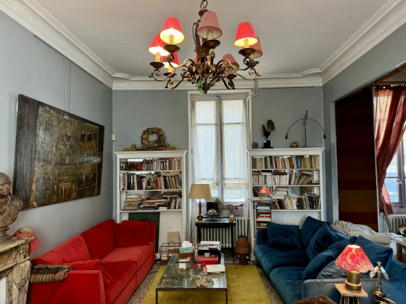Cozy living room with red and blue sofas facing each other, glass coffee table, and tall white bookshelves filled with books between two windows. A decorative chandelier hangs from the ceiling.