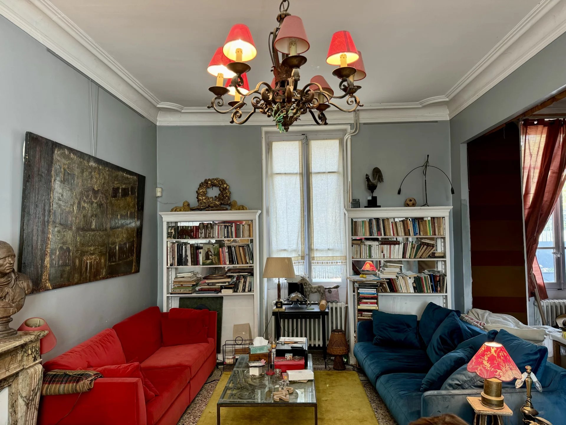 Cozy living room with red and blue sofas facing each other, glass coffee table, and tall white bookshelves filled with books between two windows. A decorative chandelier hangs from the ceiling.