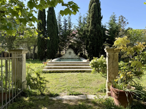 Stone fountain with a tiered basin at the end of a short set of steps in a sunny garden, flanked by tall cypress trees and green shrubbery.