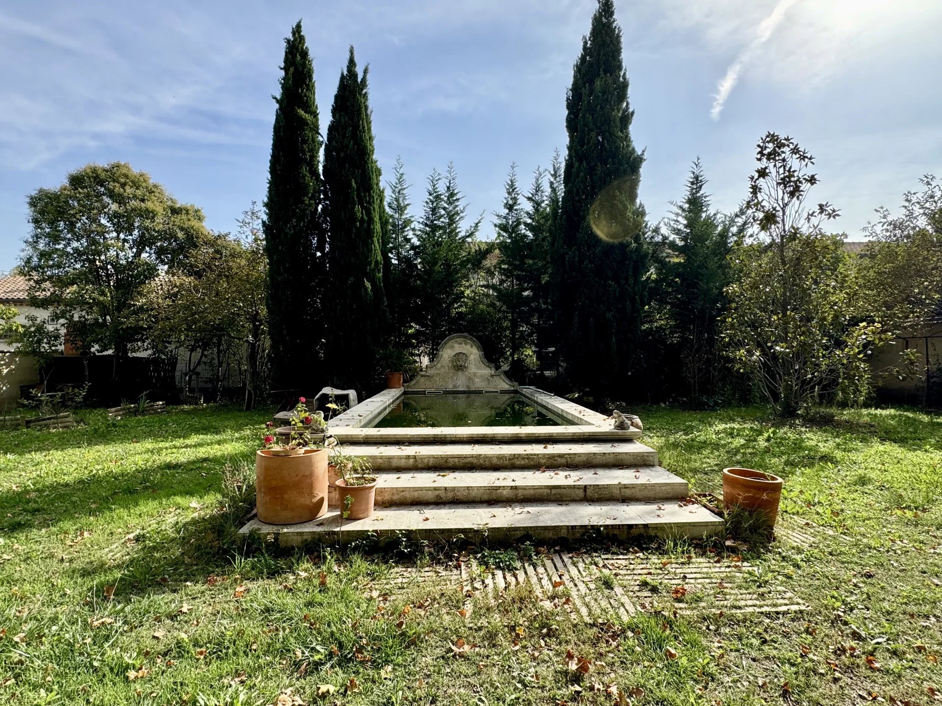 Stone fountain with stepped platform in a sunny garden, flanked by tall cypress trees and potted plants.