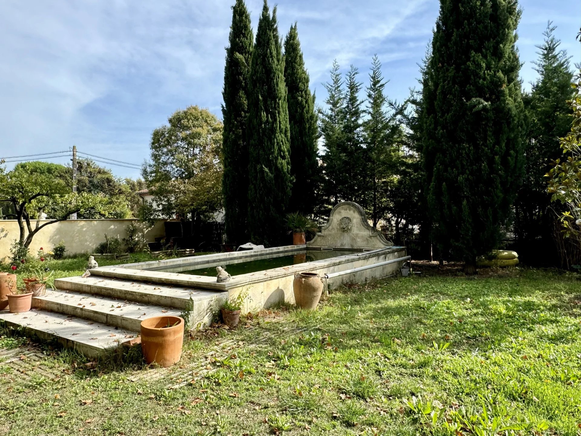 Stone rectangular fountain with steps leading to a basin, surrounded by potted plants in a sunny garden.