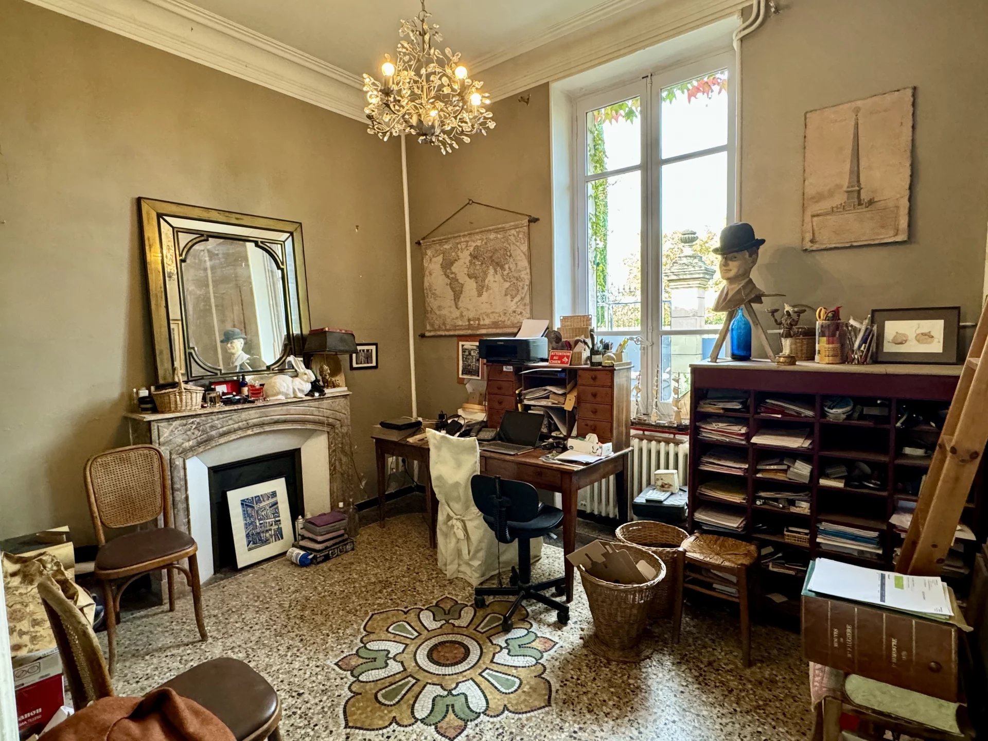 Vintage cluttered home office with a chandelier, tall window, map on wall, and a bust wearing a hat atop a desk.