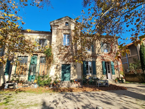 Two-story beige villa with green shutters, ivy on walls, potted plants, and a blue sky framed by autumn branches.