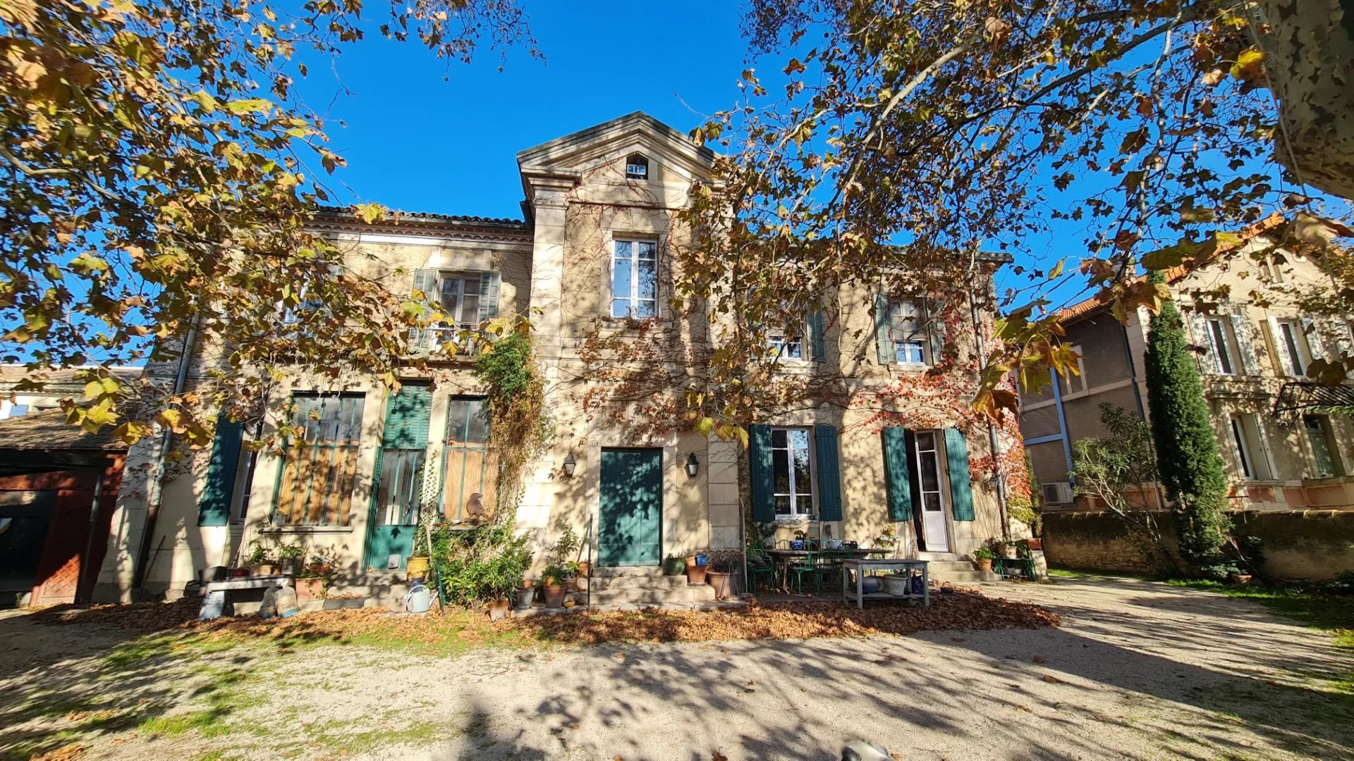 Two-story beige villa with green shutters, ivy on walls, potted plants, and a blue sky framed by autumn branches.
