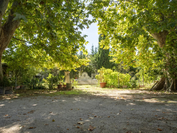 Shaded garden scene framed by large trees, with a gravel path, potted plants, and a small stone wall feature at the end surrounded by greenery.