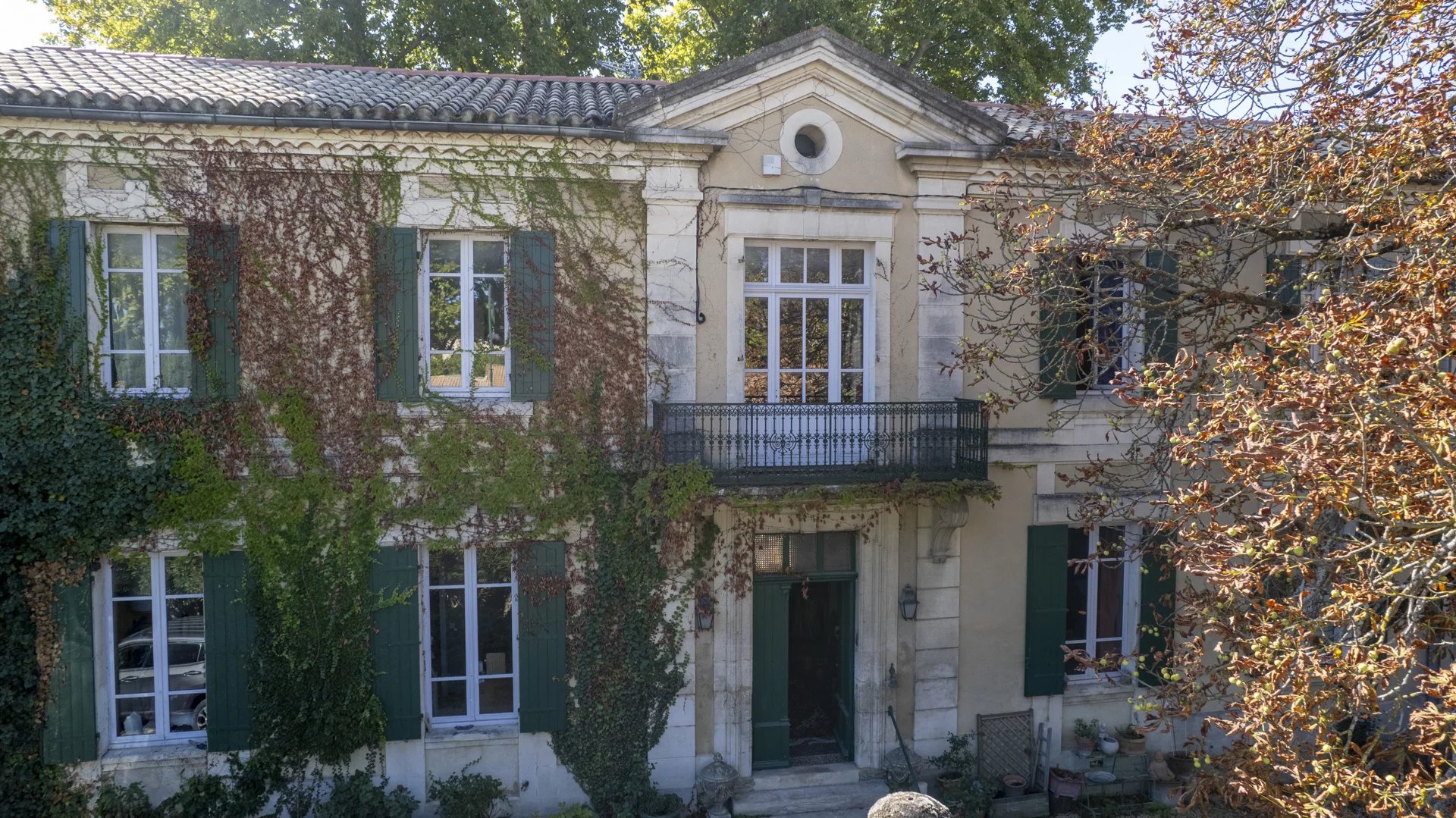 Beige two-story house with green shutters, ivy covering the walls, and a small balcony above the entrance.