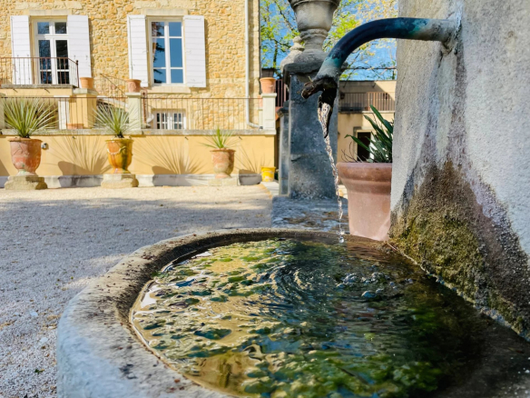 Stone fountain with flowing water, in front of a Provençal country house in the background.