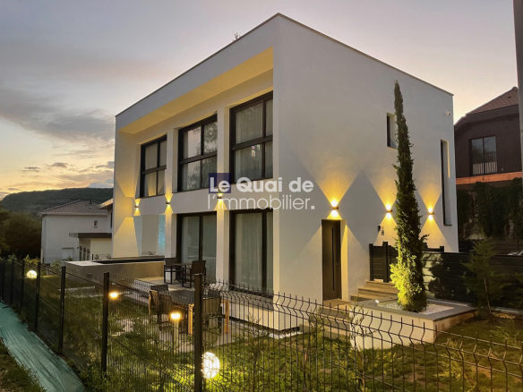 Modern white two-story house with large windows, warm exterior lighting, and a seating area on the patio at dusk with a fenced yard.