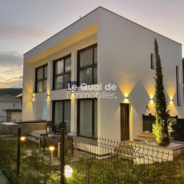 Modern white two-story house with large windows, warm exterior lighting, and a seating area on the patio at dusk with a fenced yard.