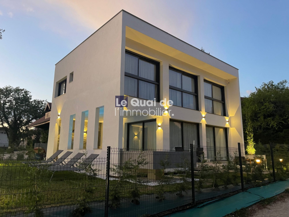 Modern white two‑story home with large black-framed windows, warm exterior lighting, and a fenced yard (watermark: Le Quai de l'immobilier).