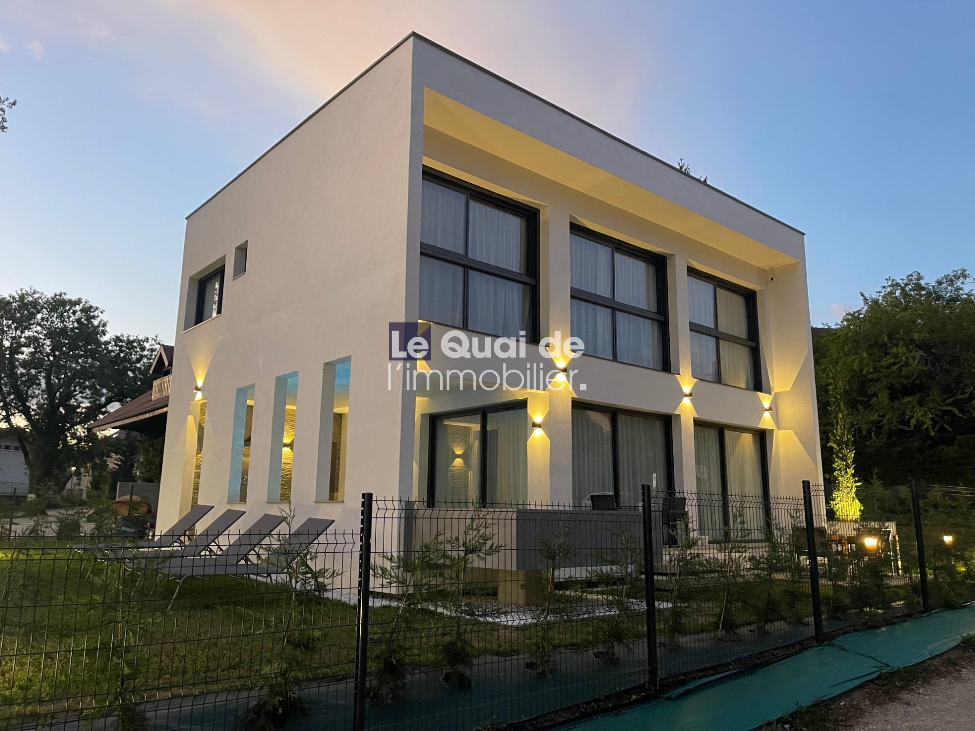 Modern white two‑story home with large black-framed windows, warm exterior lighting, and a fenced yard (watermark: Le Quai de l'immobilier).