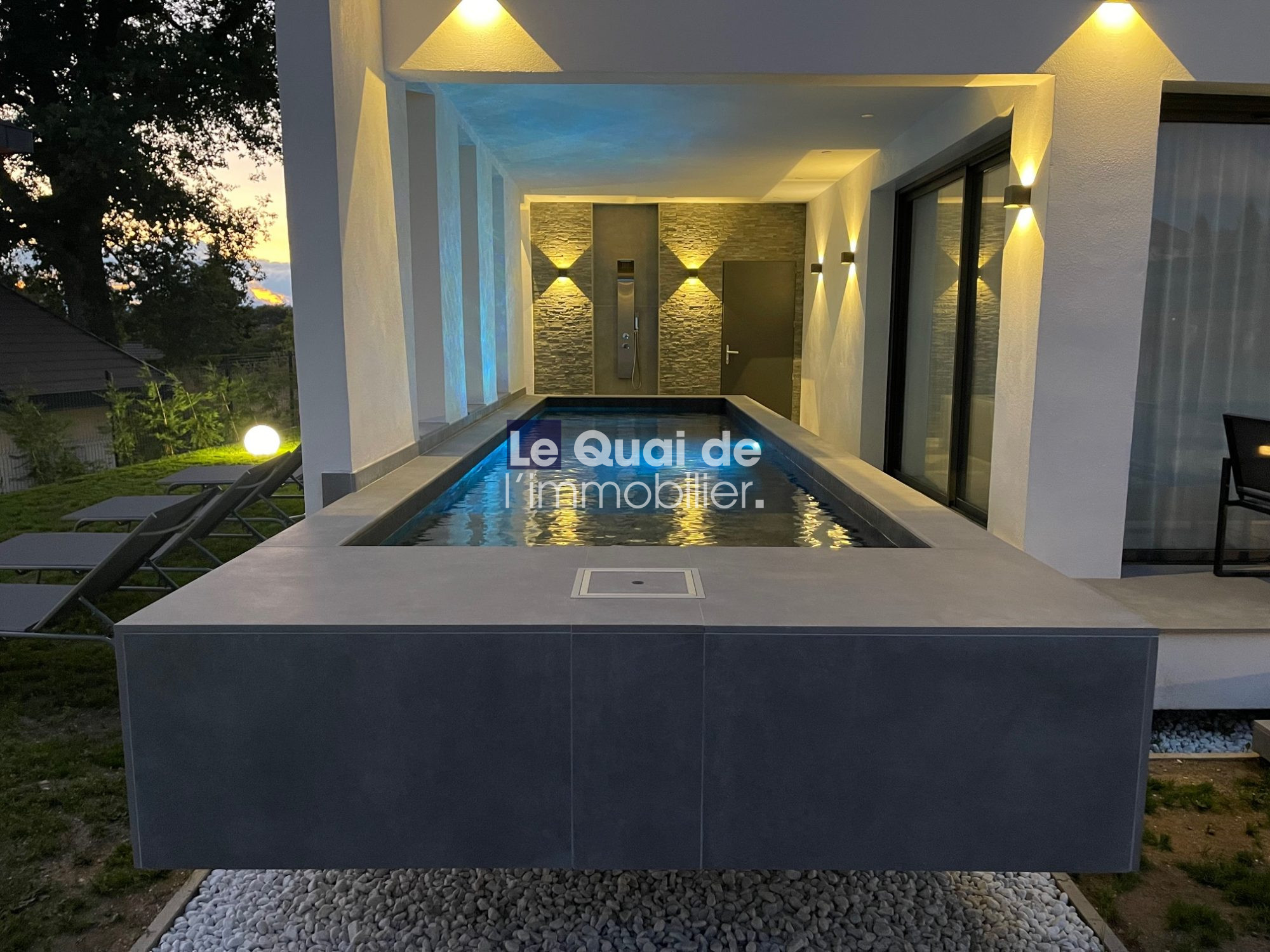 Modern rectangular pool on a covered terrace with lounge chairs and warm wall lights at dusk, reflecting the surroundings.