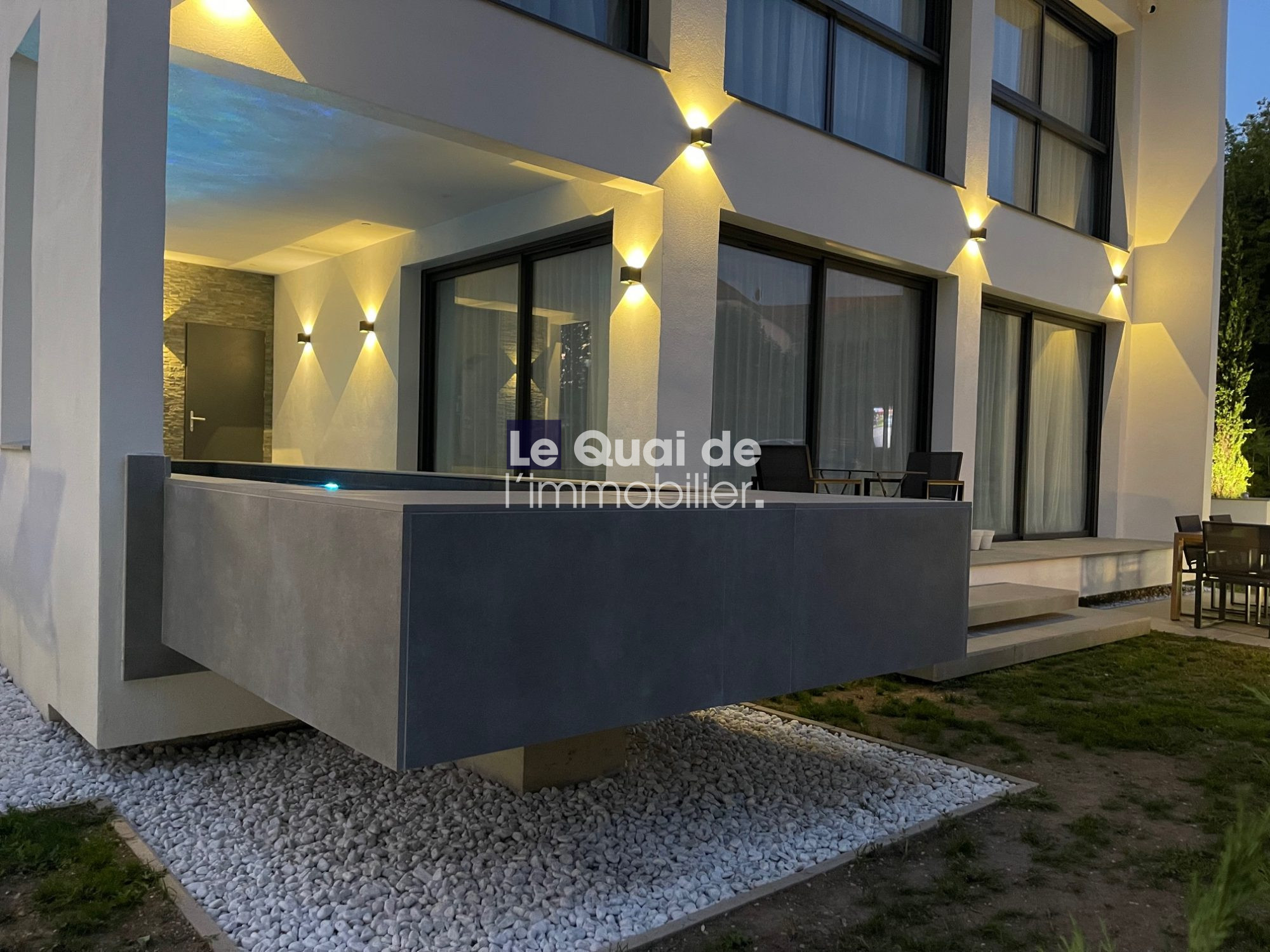 Modern house exterior at dusk with a gray cantilevered balcony, large glass doors, and warm wall lights on the walls.