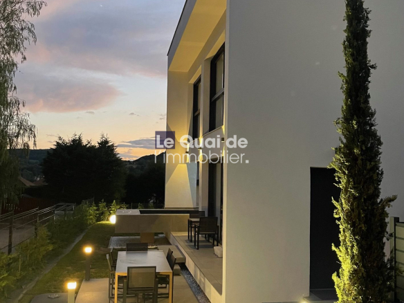 Modern white two‑story house with a lit patio, outdoor dining table and chairs on a tiled terrace, a grassy lawn with pathway lights, and a tall slender tree by the entrance.
